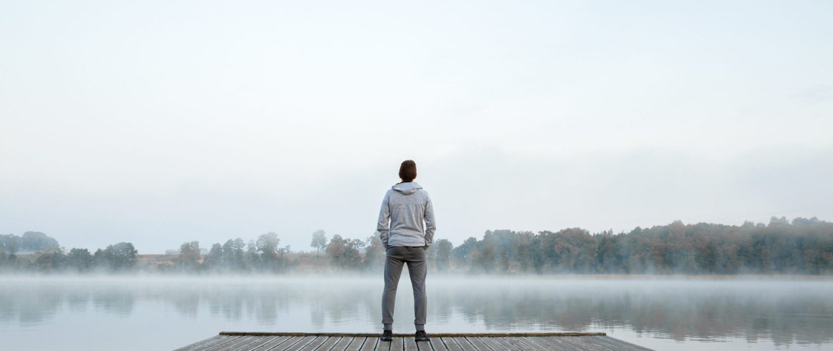 Young man standing alone on wooden footbridge and staring at lake. Thinking about life. Mist over water. Foggy air. Early chilly morning. Peaceful atmosphere in nature. Enjoying fresh air. Back view.